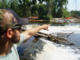 Aaron Schiller, fisheries biologist with Wisconsin Department of Natural Resources, points across the Kletzsch Dam in August 2023 toward the excavation of the Kletzsch fishway. It’s a bypass designed to unlock access to critical spawning habitat for fish like northern pike and lake sturgeon, a species once extirpated from the Milwaukee River but now being reintroduced in hopes to reestablish a naturally reproducing population.