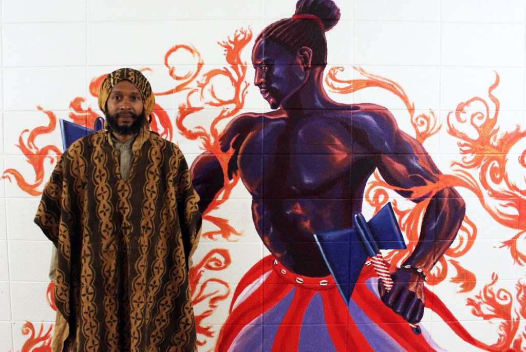 Artist Ammar Nsoroma stands with his mural painting of Shango, an Ife religion. Photo by Elizabeth Baker.