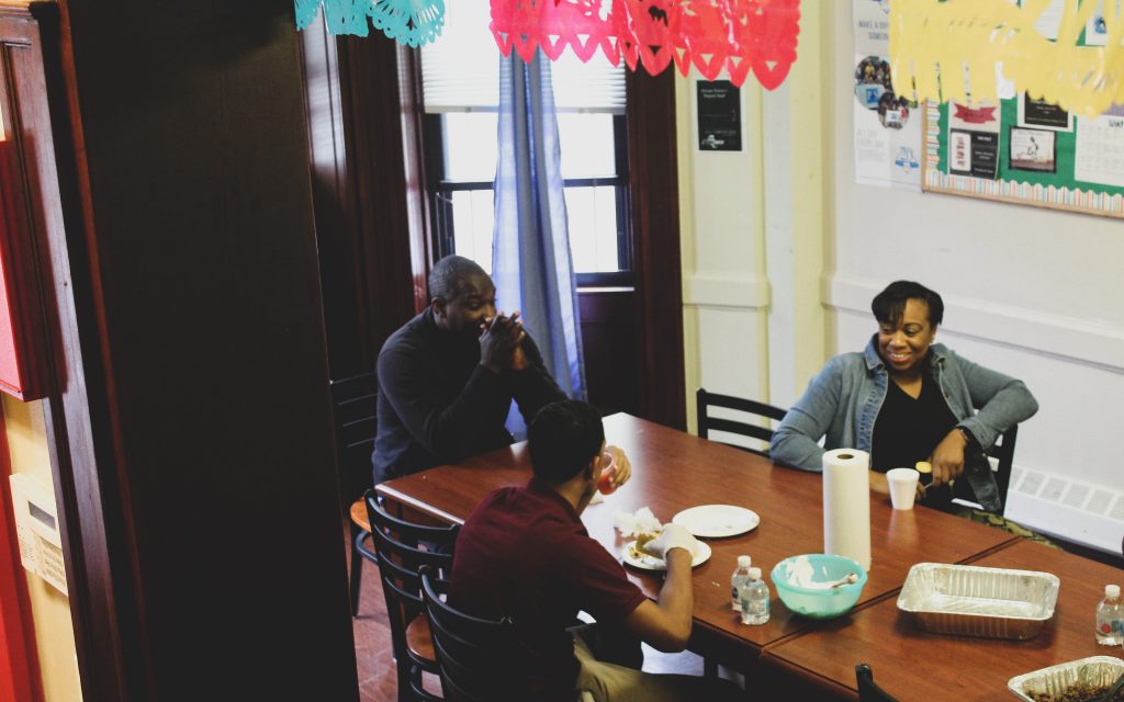 Youths eat lunch with staff members at a big family-style kitchen table at a limited-secure Close to Home facility in Brooklyn, New York. Photo by Allison Dikanovic/NNS.