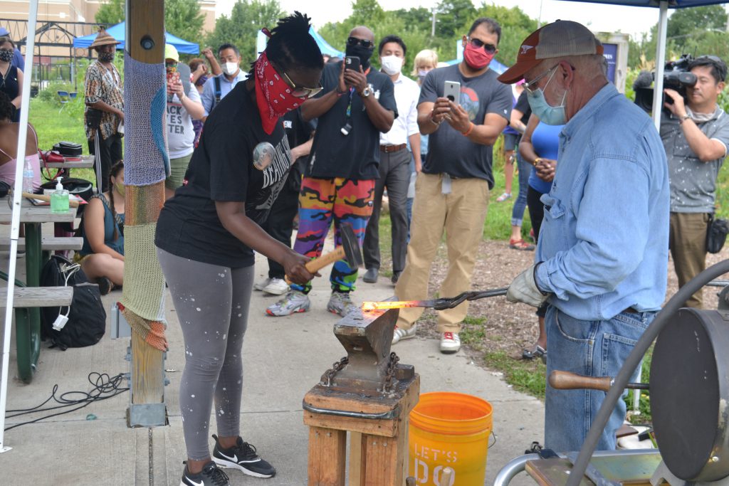 Camille Mays hammers on a piece of a gun barrel being remade into a garden tool, as Fred Martin holds it with tongs on a blacksmithing anvil. Photo by Erik Gunn/Wisconsin Examiner.