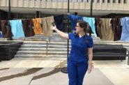 UW Health emergency room nurse Mariah Clark gestures at a line of hospital scrubs pinned up outside the City-County Building in Madison on Sept. 2, 2021. The empty hospital uniforms are mean to represent unfilled positions because of a statewide nursing shortage. The 50 scrubs each represent 200 positions. A state workforce development report projects Wisconsin will need 10,000 nurses by the end of the decade. Shamane Mills/WPR