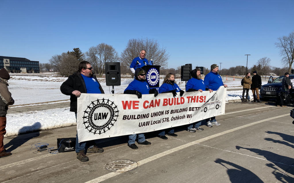 UAW Local 578 President Bob Lynk addresses a rally at Oshkosh Corp. calling on the company to build new postal trucks in Wisconsin instead of a nonunion plant in South Carolina. Members of the local’s bargaining committee stand in front. Photo by Miles Maguire/Wisconsin Examiner.