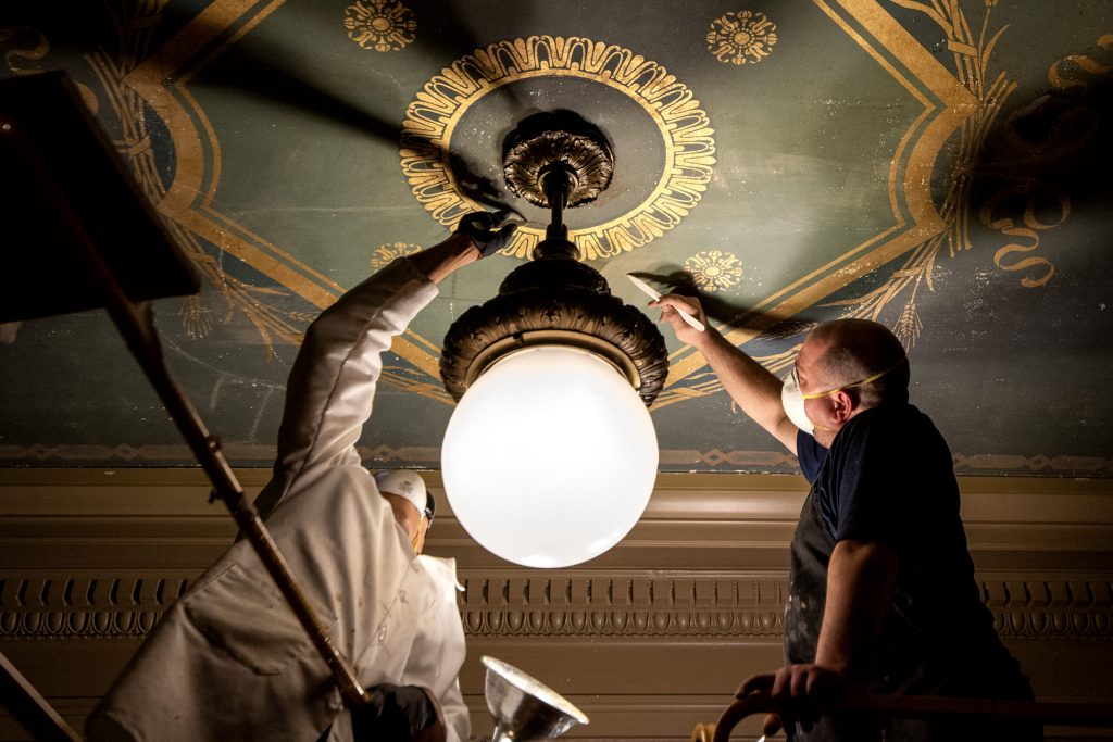 Conservation technicians Tim Phebus, right, and Jose Anico, left, use a bone folder to remove yellowing varnish installed in the 1960’s on Thursday, Dec. 14, 2023, at the Wisconsin State Capitol in Madison, Wis. Angela Major/WPR
