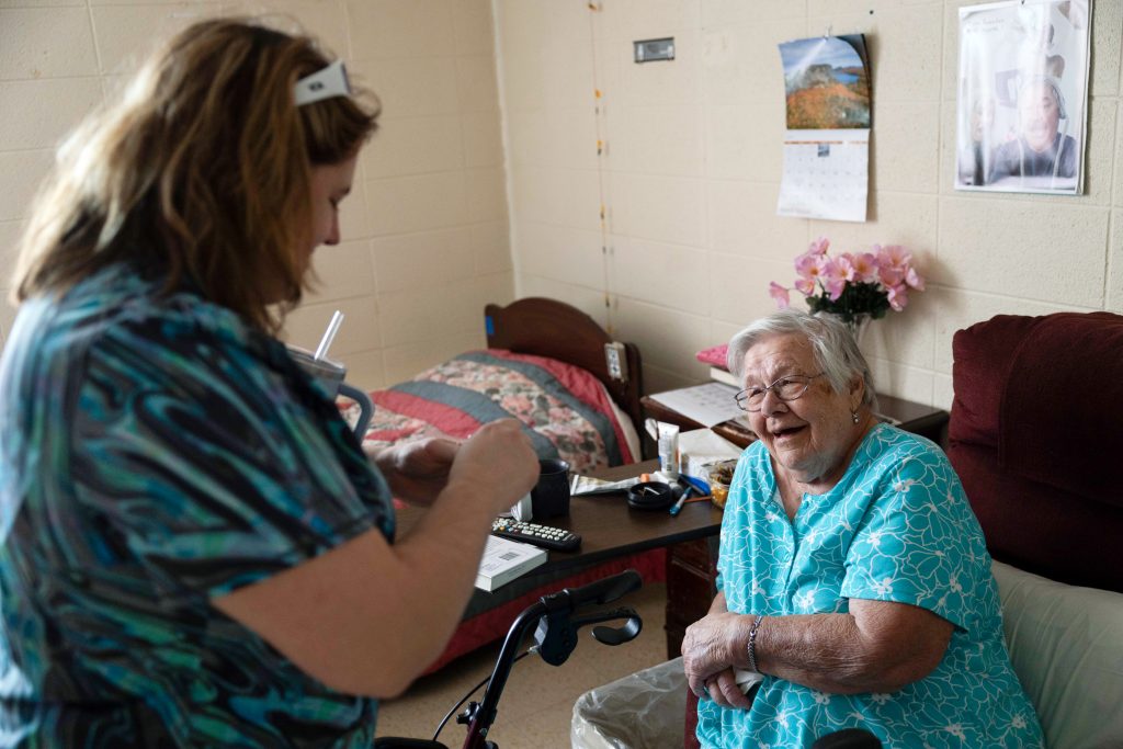 Arlene Meyer, 86, of Gleason, right, smiles as Paula Streich, a certified nursing assistant, left, opens a chocolate bar she brought for Meyer on Nov. 15, 2024, at Pine Crest Nursing Home in Merrill, Wis. Meyer said Streich often brings her gifts from outside the nursing home like chocolate bars. (Joe Timmerman / Wisconsin Watch)