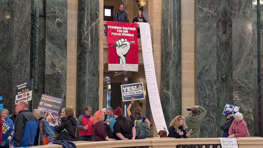 A rally goer rolls out a scroll with the names of every school district that has gone to referendum since the last state budget. Photo by Baylor Spears/Wisconsin Examiner.