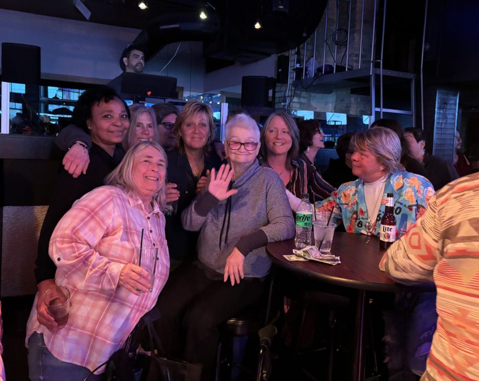Sharon Dixon waves at the camera, surrounded by old friends and patrons of her past LGBTQ+ women’s bars, at a celebration in her honor on March 30, 2025 at La Cage nightclub in Milwaukee. Beatrice Lawrence/WPR