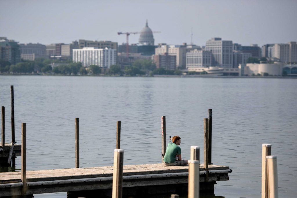 A man sits on a dock as smoke shades the view of the Wisconsin State Capitol across Lake Monona on Wednesday, June 4, 2025, in Madison, Wis. Angela Major/WPR