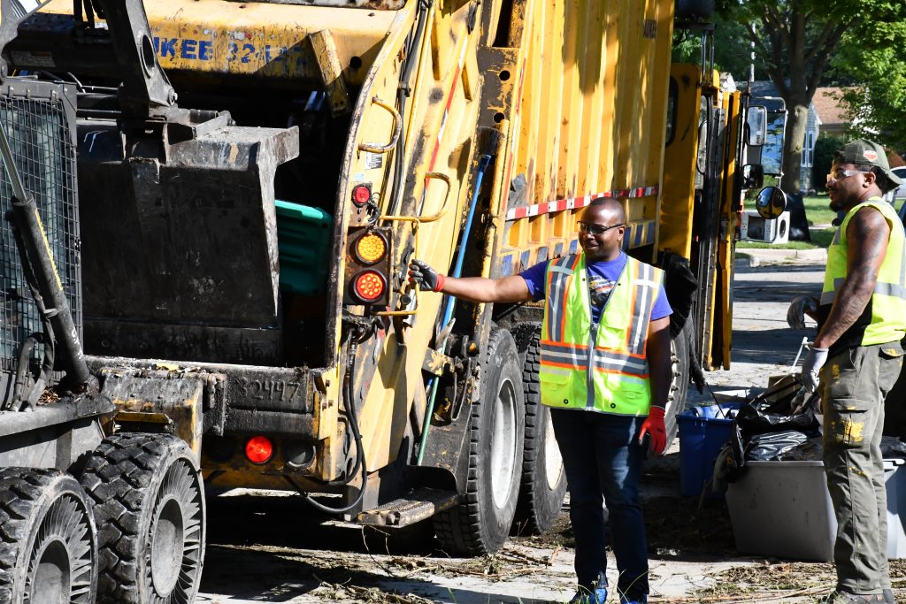 Mayor Cavalier Johnson holds onto a city garbage truck while a skid steer deposits flood debris. Photo by Jeramey Jannene.