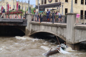 People gather near the bridges in the Wauwatosa village to observe the still rushing flooded river and storm damage. (Photo by Isiah Holmes/Wisconsin Examiner)