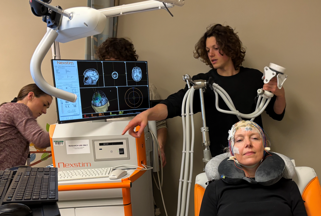 UW-Madison researchers in professor Charles Raison’s lab study how psilocybin works in the brain. Here, study team members Emma Bublitz (left), James Barlow (behind screen), Michael Sutherland (center right), and Laura McCormick (in chair) set up an electroencephalogram to measure her brain’s electrical activity. Photo courtesy of the Raison Lab