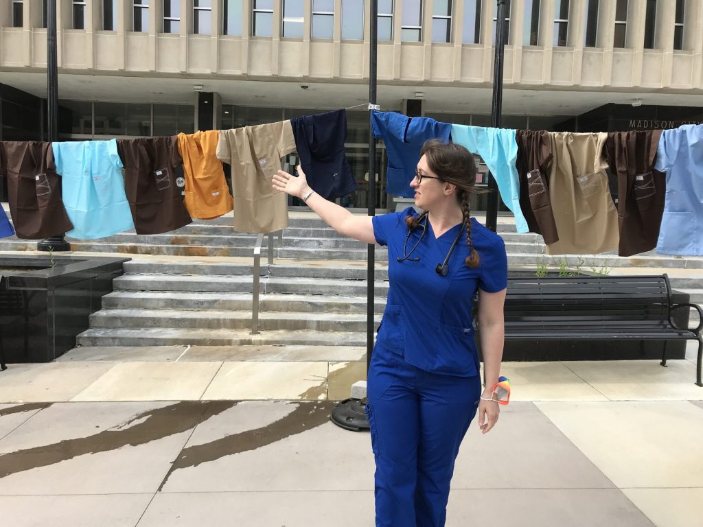 UW Health emergency room nurse Mariah Clark gestures at a line of hospital scrubs pinned up outside the City-County Building in Madison on Sept. 2, 2021. The empty hospital uniforms are mean to represent unfilled positions because of a statewide nursing shortage. The 50 scrubs each represent 200 positions. A state workforce development report projects Wisconsin will need 10,000 nurses by the end of the decade. Shamane Mills/WPR