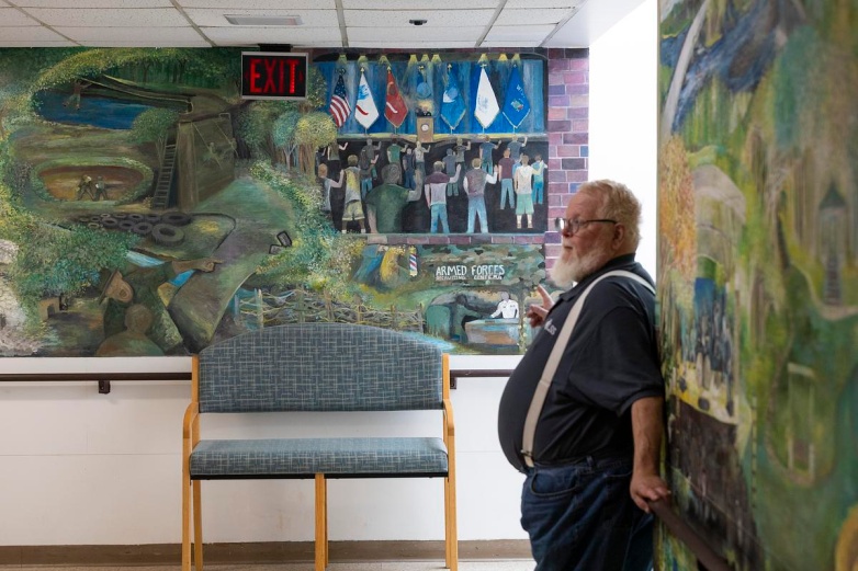 Randy Withrow, site director of Klein Hall, stands in front of a mural painted by veteran James Heber that depicts Chippewa Falls and a veteran’s journey at Klein Hall is painted on the wall, Monday, Aug. 25, 2025, at Klein Hall in Chippewa Falls, Wis. (Joe Timmerman / Wisconsin Watch)