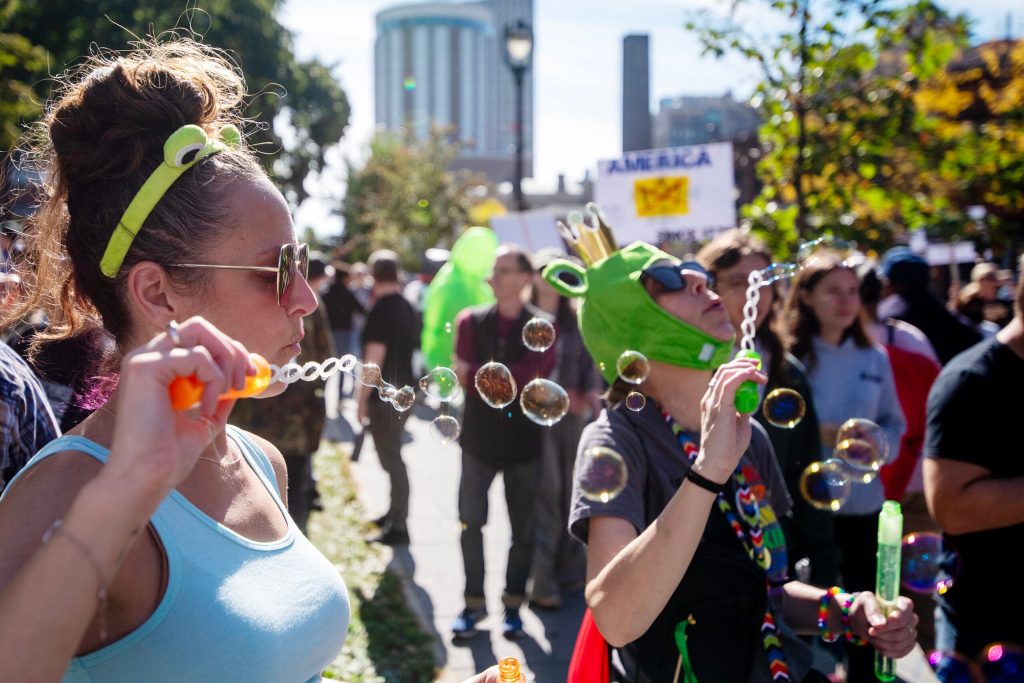 Protesters Sarah Young, left, and Angie Lawrence, right, wear frog hats and blow bubbles during an anti-Trump protest Saturday, Oct. 18, 2025, at Cathedral Square Park in Milwaukee, Wis. Angela Major/WPR