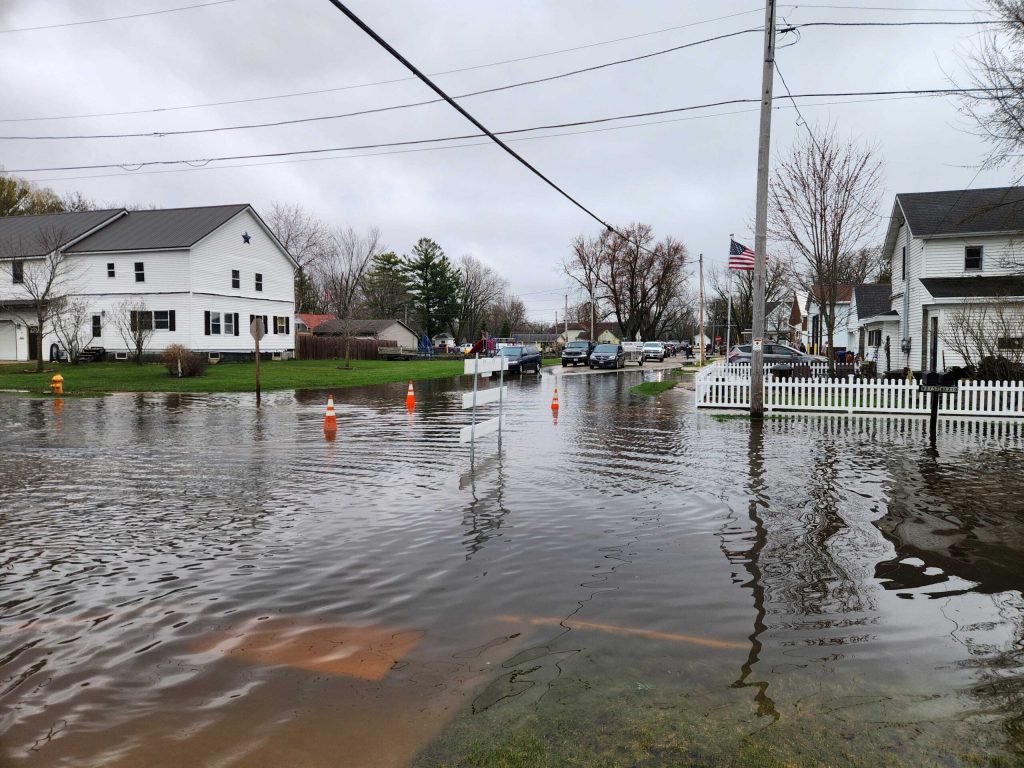 A flooded residential street is seen with water covering the road and lawns, traffic cones blocking access, and overcast sky in Shiocton, Wis., on April 15, 2026. Joe Schulz/WPR