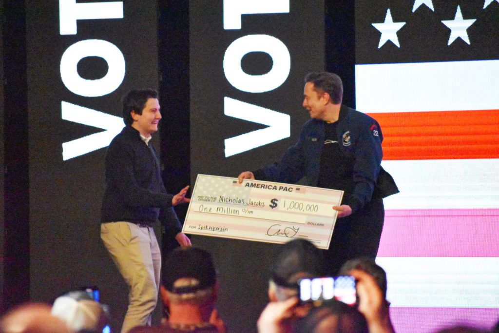 Elon Musk, right, hands a check to Nicholas Jacobs, president of the Wisconsin College Republicans, during a town hall in Green Bay on Sunday, May 30, 2025. Joe Schulz/WPR