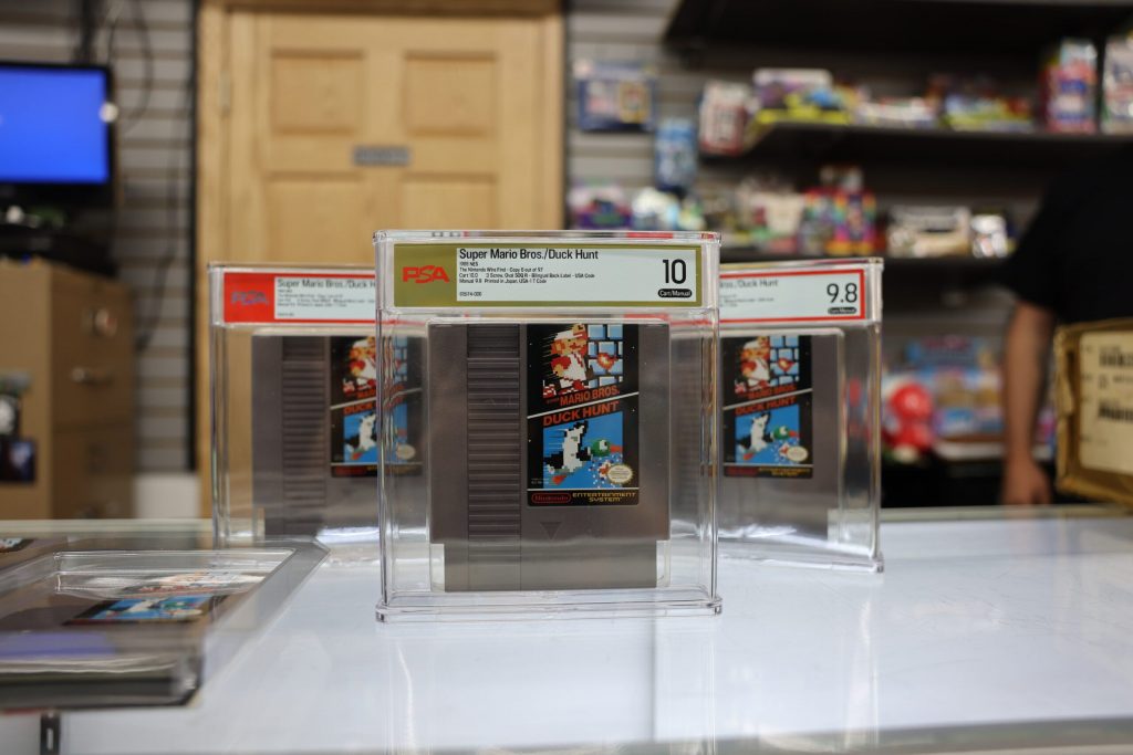 Three graded copies of the Super Mario Brothers Duck Hunt Nintendo Entertainment cartridge sit on the counter of One Stop Wonders video game and collections shop in Waukesha on Aug 12, 2026. The center copy is graded a 10, meaning it is in perfect condition. This is the first NES cartridge to receive a perfect grade from the Professional Sports Authority. Elissa Eaton/WPR