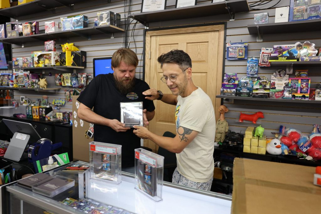Kevin Braun, left, and Jason Ganos, right, hold a cartridge of Super Mario Brothers Duck Hunt at One Stop Wonders, a video game and collections shop in Waukesha on Aug. 12, 2026. Elissa Eaton/WPR