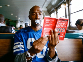 A grown man reads at an indeterminate location with a pacifier in his mouth. The book is entitled “Laugh-Out-Loud Jokes For Kids.” No kids are in sight. The man appears unamused.