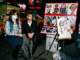 A couple sits for a caricaturist. The woman wears a surgical mask, but not in the nearly finished drawing.